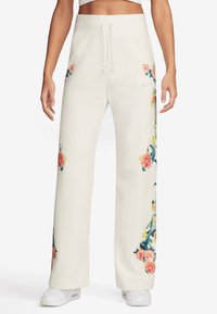 Kvinna som bär lösa vita byxor med rosa och gula blommönster på sidorna, i kombination med vita sneakers och en vit cropped topp.