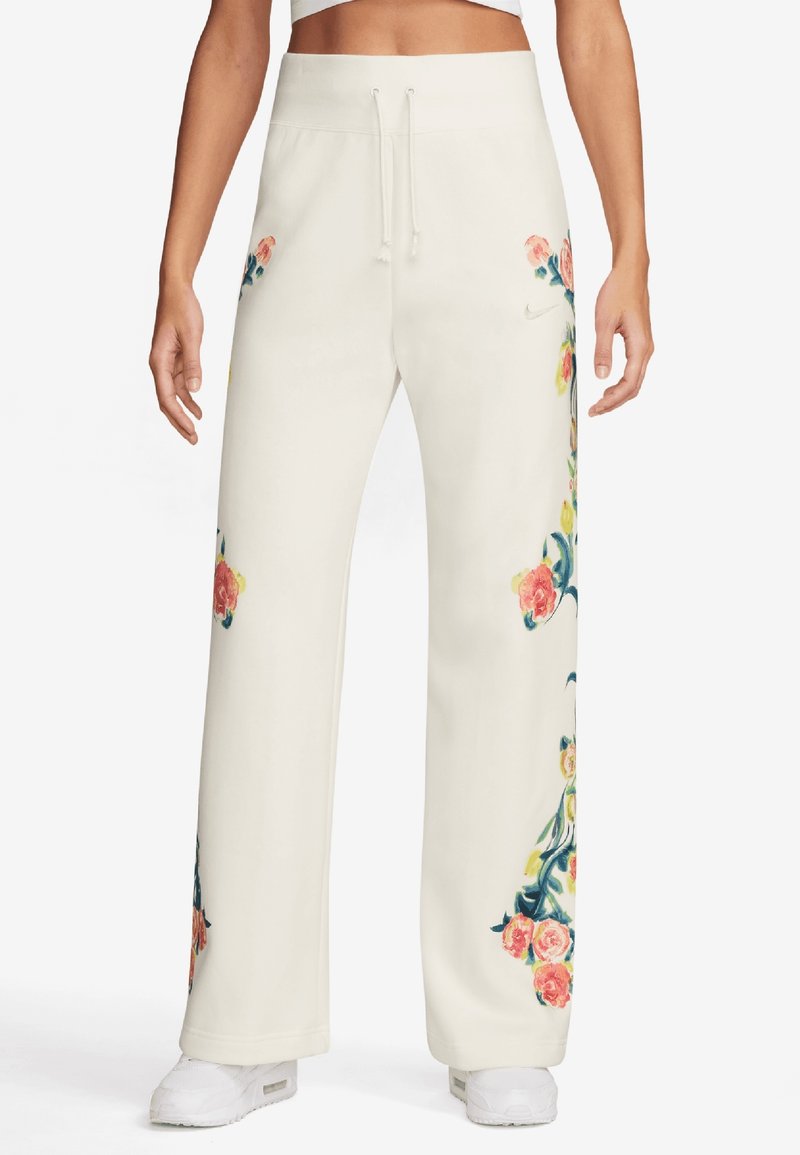 Kvinna som bär lösa vita byxor med rosa och gula blommönster på sidorna, i kombination med vita sneakers och en vit cropped topp.