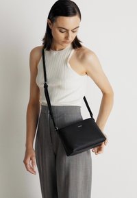 Svart läder crossbody-väska med rektangulär form, justerbar rem och subtilt logo på framsidan. Slät textur, minimalistisk design.