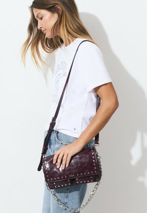 Joven con cabello largo que lleva una camiseta blanca estampada y jeans de mezclilla azul claro, sosteniendo un bolso cruzado de cuero burdeos oscuro con tachuelas de metal.