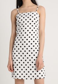 Robe blanche à pois noirs, avec fines bretelles et corsage ajusté. Le tissu semble léger et légèrement texturé.