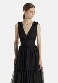 Robe noire avec un décolleté en V profond, jupe en tulle superposée, taille ajustée et accents transparents. Tissu léger avec une silhouette fluide.