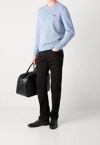 Maglione azzurro chiaro con un piccolo logo rosso, pantaloni neri e borsa in pelle nera. Le scarpe sono in pelle scura con una finitura liscia.