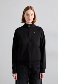 Crna zip-up dukserica s visokim ovratnikom, s zlatnim logotipom, raglan rukavima i prednjim džepovima. Glatka tekstura i usklađen dizajn.