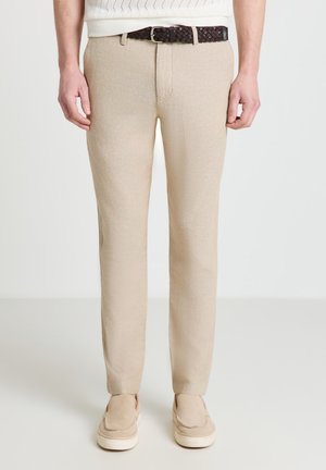 Homme portant un pantalon beige slim, une ceinture tissée sombre et des chaussures beiges à enfiler assorties sur un fond uni.