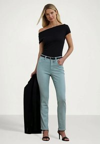 Moteris vilki juodą off-shoulder viršų, šviesiai mėlynus džinsus su juodu diržu, juodas aukštakulnius batus, o vienoje rankoje laiko juodą švarką.