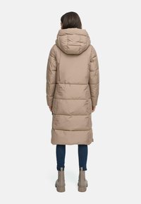 Beige pufferjakke med hætte, der har vandret syninger, lang længde og glat tekstur. Iført med blå jeans og lyse støvler.