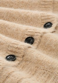 Beżowa dzianina o miękkiej fakturze, z trzema czarnymi guzikami na przodzie. Projekt zawiera wypukłe wzory dla dodania detali.