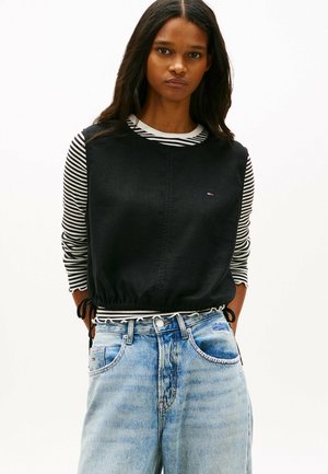 Vrouw met lang donker haar die een zwart mouwloos top draagt over een zwart-wit gestreept shirt met lange mouwen en lichtblauwe hoge taille jeans.