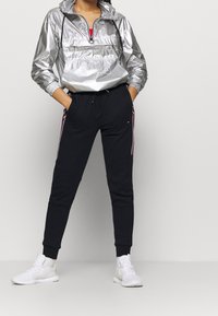 Chaqueta ligera plateada con capucha y media cremallera, combinada con pantalones jogger negros que tienen rayas laterales rojas y blancas. Zapatillas blancas.