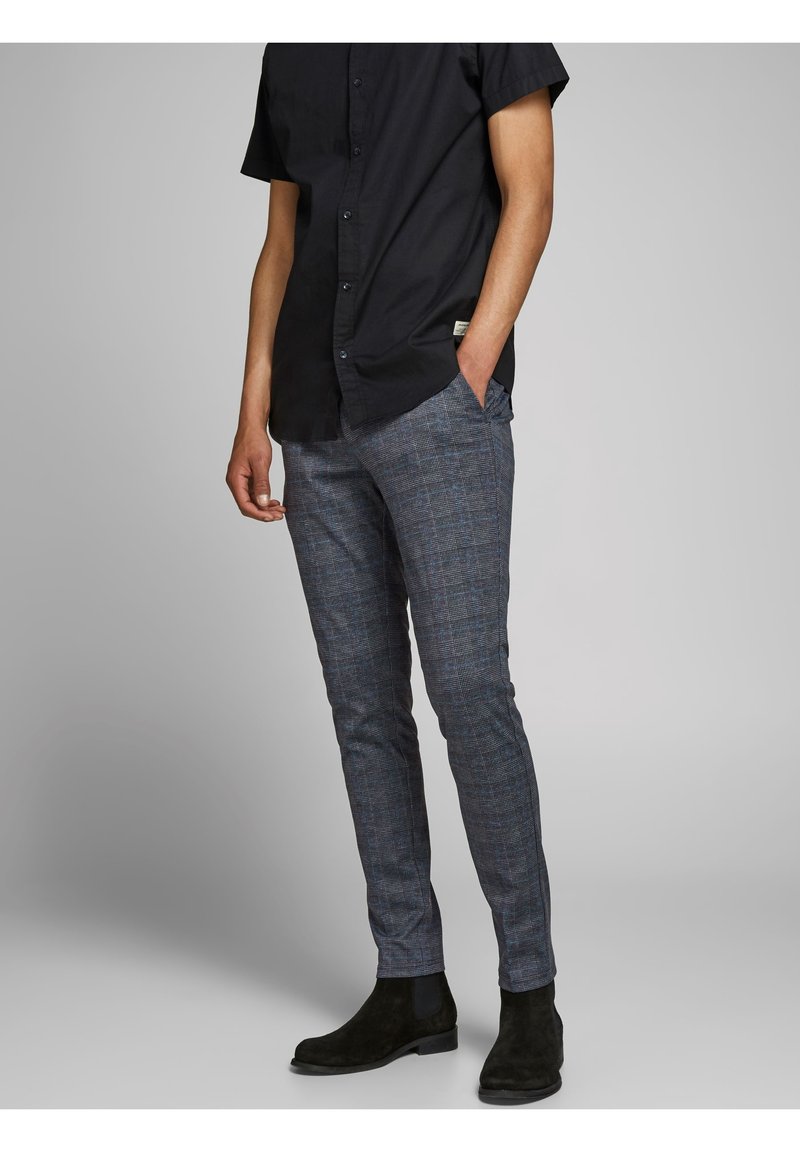 Homme portant un pantalon gris à carreaux slim, une chemise noire boutonnée et des bottines en daim noir, debout avec une main dans la poche.