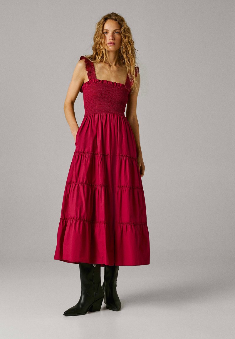 Moteris su banguotais plaukais, vilkinti raudoną be rankovių sluoksniuotą midi suknelę ir juodus vidutinio aukščio aulinukus, stovinti priešais paprastą foną.