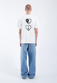 Camiseta blanca con diseños de un corazón negro y un símbolo de paz en la parte posterior, combinada con un par de jeans de mezclilla claro de corte holgado.