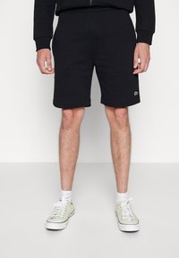 Svarta bomullsshorts med elastisk midja, sydda fållar och en liten grön logotyp på vänster lår. Bärs med ljusgröna sneakers.
