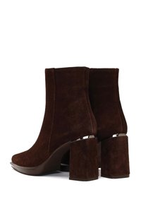 Botines de ante marrón con un diseño elegante, con un tacón cuadrado grueso y sutiles detalles plateados en el tacón. Superficie texturizada.