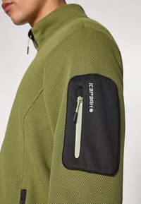 Πράσινο σακάκι από δίχτυ με μαύρη τσέπη με φερμουάρ στο πάνω μέρος του βραχίονα. Η υφή είναι σαν κυψέλη, με ψηλό γιακά και διακριτικό λογότυπο.