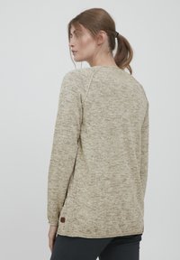 Pull à manches longues beige avec un design texturé et chiné. Caractéristiques des manches raglan et une petite étiquette brune en bas à gauche.