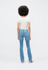 Persoană cu păr afro creț, purtând un top mulat de culoare alb murdar, blugi de denim albastru deschis și pantofi albi cu toc, stând cu spatele către privitor.