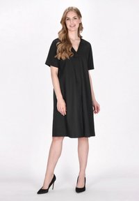 Schwarzes Kleid mit V-Ausschnitt, kurzen Ärmeln und lockerer Passform. Glatter Stoff und knielanger Saum. Getragen mit schwarzen High Heels.