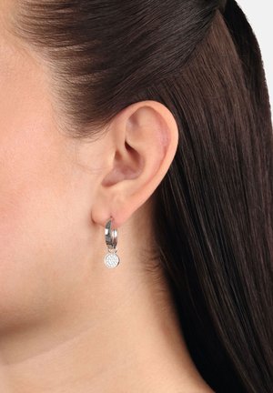 Kleine silberne Creole mit einem runden, mit Kristallen besetzten Charme, die am linken Ohr einer Person mit dunkelbraunem Haar getragen wird.