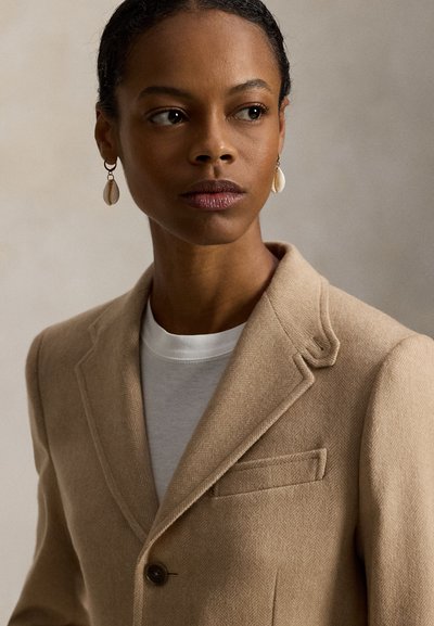 Béžinis kailiniai švarkas su įpjauta apykakle, vieno mygtuko užsegimu ir krūtinės kišene dėvimas per baltą marškinėlį; derinamas su kriauklės formos auskarais.