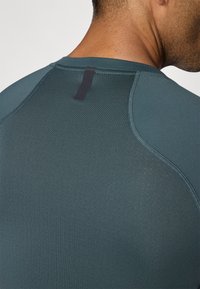 T-shirt de sport en bleu sarcelle foncé avec une texture lisse, doté d'un col court et d'une étiquette noire en boucle à l'arrière du cou. Détails en mesh sur les épaules.