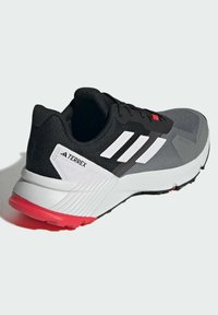 Zapatilla de running Adidas Terrex en negro y gris con rayas blancas y suela rosa, vista lateral en ángulo sobre fondo claro.