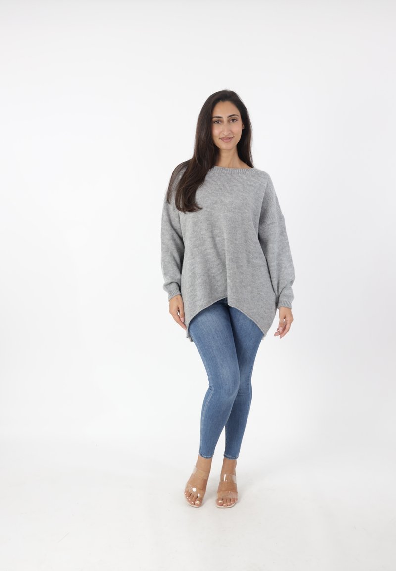 Grijze oversized trui met een wijde halslijn en asymmetrische zoom, gecombineerd met blauwe skinny jeans en doorzichtige sandalen op een witte achtergrond.