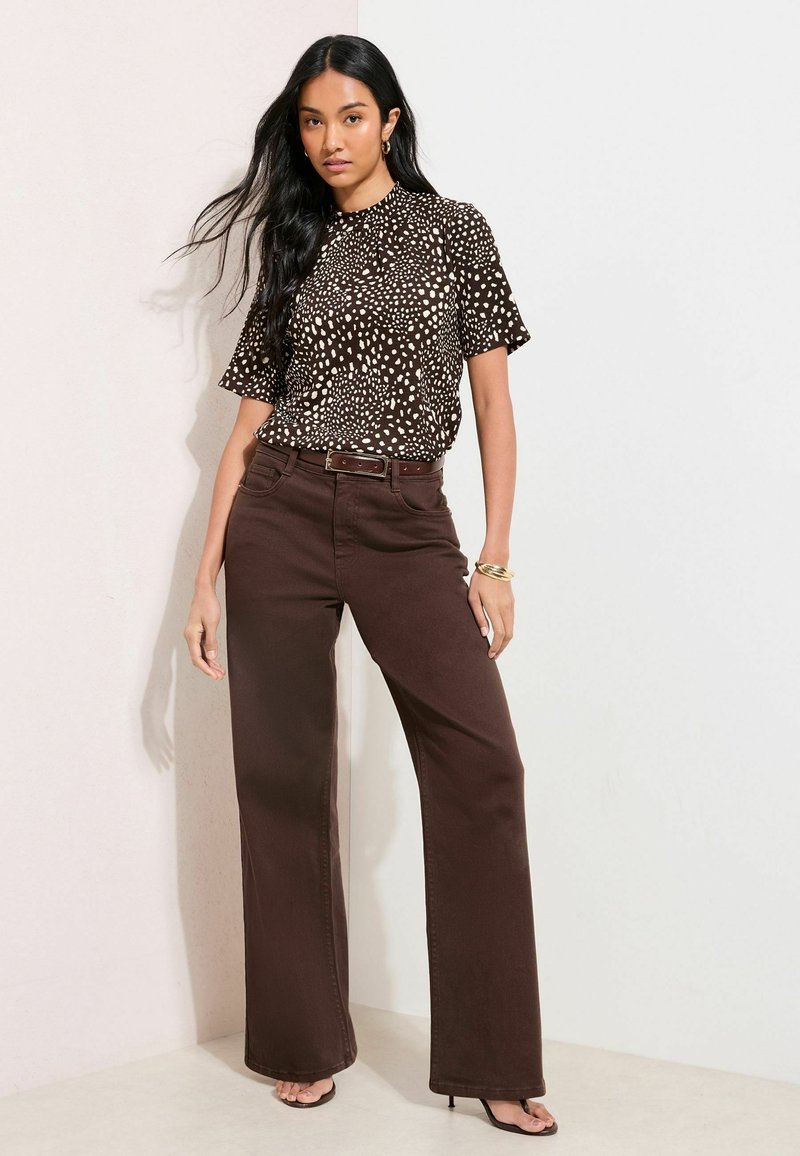 Top a maniche corte marrone maculato abbinato a pantaloni larghi a vita alta marroni. Orecchini a cerchio dorati e tacchi aperti completano l'outfit.