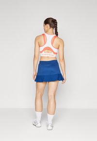 Conjunto atlético com um top desportivo branco e laranja com um design recortado e uma saia plissada azul. Meias brancas até ao tornozelo e ténis cinzentos.