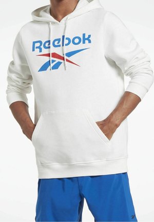 Hvid hættetrøje med blå og rød Reebok-logo, frontlomme, båret af person med hænderne i lommerne, kombineret med blå shorts.