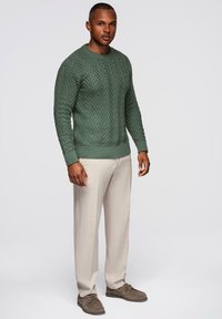 Zielony sweter z dzianiny o wzorze zygzakowym, okrągłym dekolcie i ściągaczami na rękawach. Połączony z jasnobeżowymi spodniami i brązowymi butami.