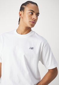 Camiseta branca de algodão com decote redondo e mangas curtas, apresentando o logótipo cinza da New Balance na área do peito esquerdo.