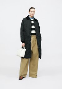 Długa czarna pikowana kurtka, sweter w paski, szerokie beżowe spodnie i biała torebka; osoba nosi granatowy kołnierz i brązowe buty.