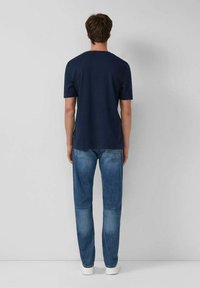 Mann steht mit dem Rücken zu einer weißen Wand, trägt ein navyblaues Kurzarm-T-Shirt, blaue Jeans und weiße Sneakers.