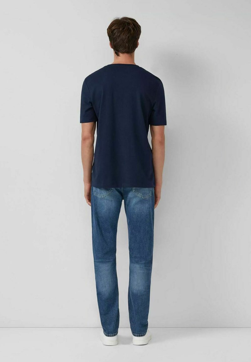 Mann steht mit dem Rücken zu einer weißen Wand, trägt ein navyblaues Kurzarm-T-Shirt, blaue Jeans und weiße Sneakers.