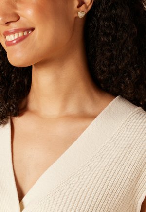 Femme souriante portant un haut en maille blanche et de petites boucles d'oreilles en forme de cœur en or avec un centre en perle et une bordure en strass.