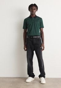 Zelená polo košeľa s čiernym golierom, skombinovaná s tmavými džínsami a bielymi teniskami. Jednoduchý dizajn s gombíkovou légou a klasickým strihom.