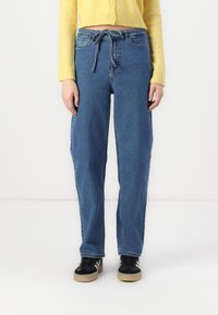 Jeans de denim azul claro com um ajuste relaxado, apresentando uma cintura com cordão e bolsos laterais, combinados com sapatos pretos com sola de borracha.