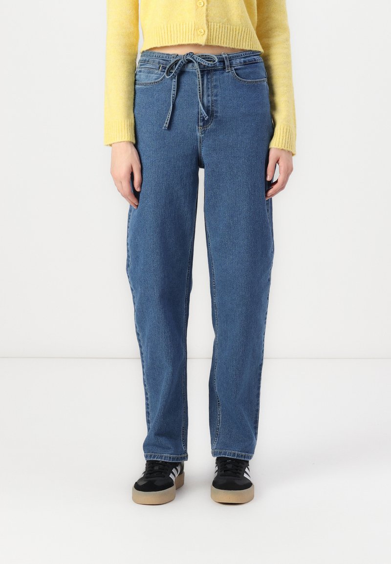 Jeans de denim azul claro com um ajuste relaxado, apresentando uma cintura com cordão e bolsos laterais, combinados com sapatos pretos com sola de borracha.