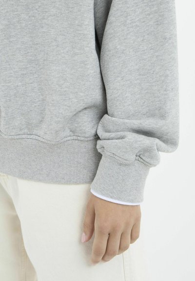 Felpa grigia con orlo e polsini a costine, caratterizzata da una morbida texture e dettagli di maniche leggermente arricciate, indossata su pantaloni bianchi.