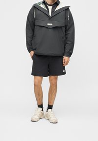 Personne portant un coupe-vent noir Columbia avec une doublure de capuche fleurie, un short noir, des chaussettes noires et des chaussures de sport beiges, debout devant un fond uni.