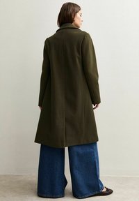 Cappotto verde oliva con design su misura, maniche lunghe e taglio a metà lunghezza, abbinato a pantaloni in denim blu a gamba larga. Texture liscia, senza motivi visibili.