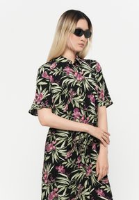 Robe florale noire avec des orchidées violettes et des feuilles vertes. Manches courtes, fermeture à boutons à l'avant et détail de taille nouée. Le modèle porte des lunettes de soleil noires.