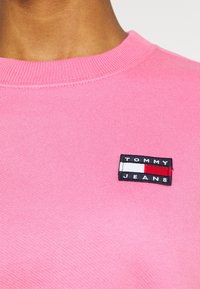 Maglione in cotone rosa con scollo rotondo a costine e un patch ricamato rettangolare con "TOMMY JEANS" in bianco e nero sul petto.