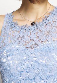 Světle modré krajkové šaty s květinovou výšivkou a průsvitnou vrstvou, pod kterou je hladká saténová podšívka. Zlatý náhrdelník s černým přívěskem.