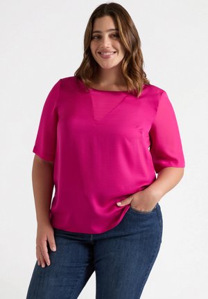 Blusa de manga corta en rosa caliente con escote cuadrado y un inserto en forma de V, combinada con jeans azules oscuros. Textura suave, ajuste casual.