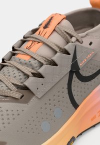 Chaussure de trail Nike beige avec semelle et languette orange ornées d'un motif d'épi de blé, lacets gris et logo Nike swoosh noir.