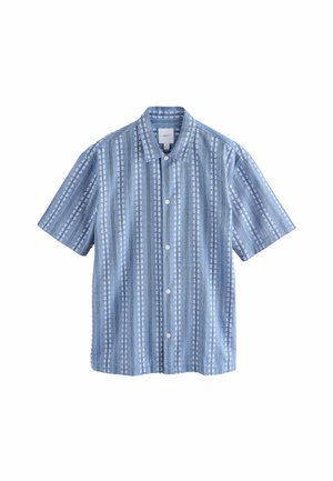 Chemise bleue à motif avec des rayures florales blanches portée sur un pantalon beige, montrant une couture latérale et une petite fente à l'ourlet de la chemise.
