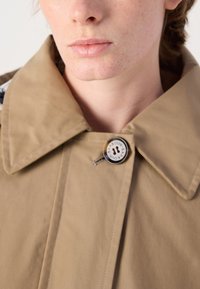 Beige Trenchcoat mit großem Kragen, ausgestattet mit einem silbernen Knopf mit eingraviertem Text. Glatte Textur und sichtbare Nähdetails.